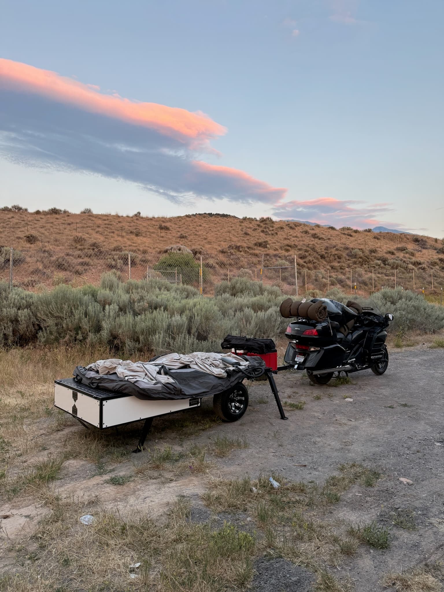 Black motorcycle with gear hitched to a white and black overland camping trailer, folded tent on top, parked in a dry, bushy field with hills and sunset clouds.
