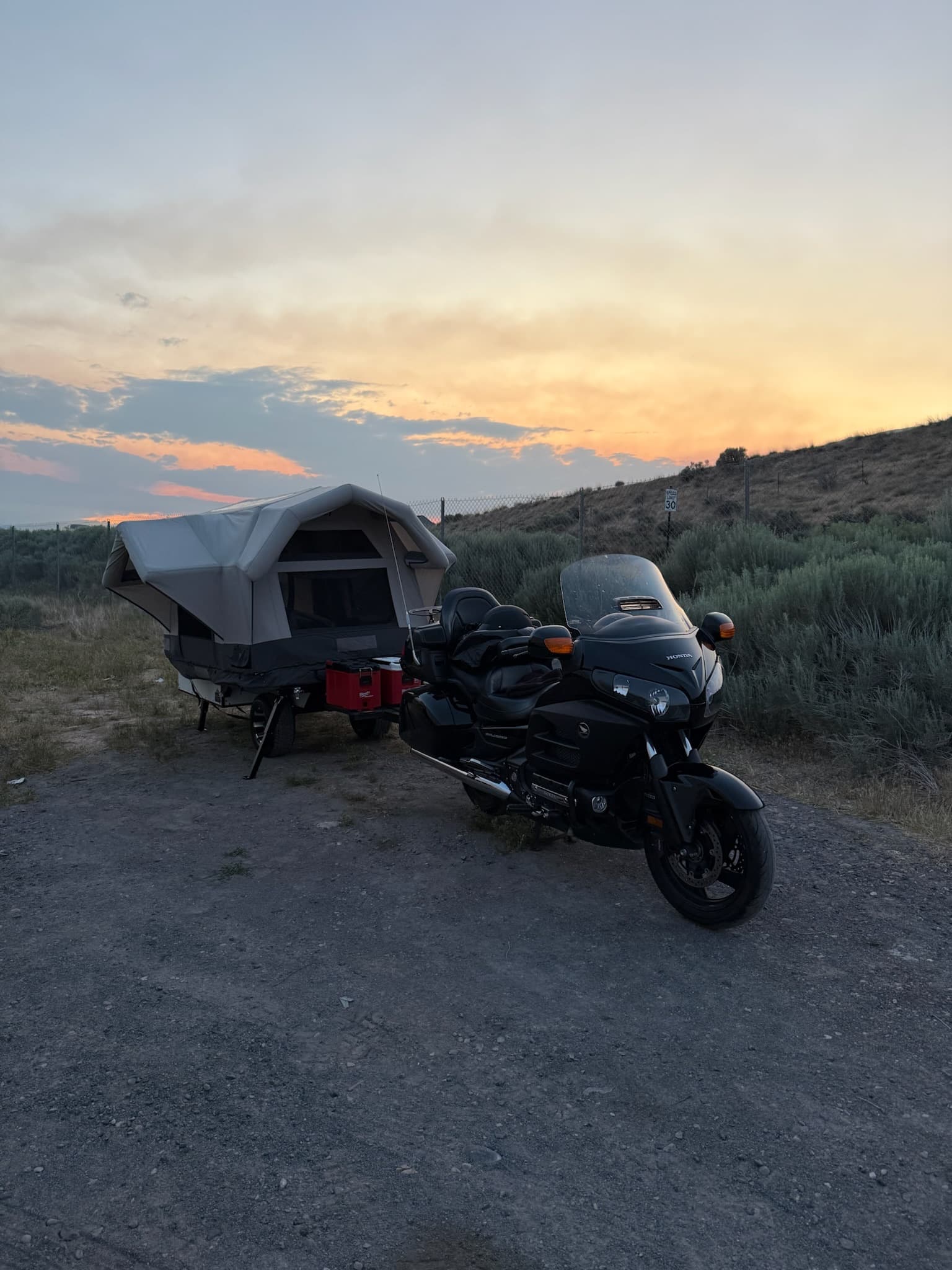 Hand-built overland camping trailer with pop-up tent and black Honda motorcycle on a gravel path at sunset, beside a brushy hillside.