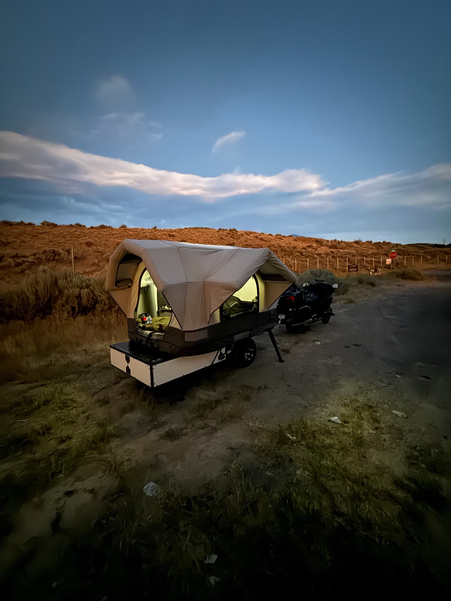 Ultra Compact Conestoga trailer with its airframe tent deployed and glowing at dusk, hitched to a touring motorcycle