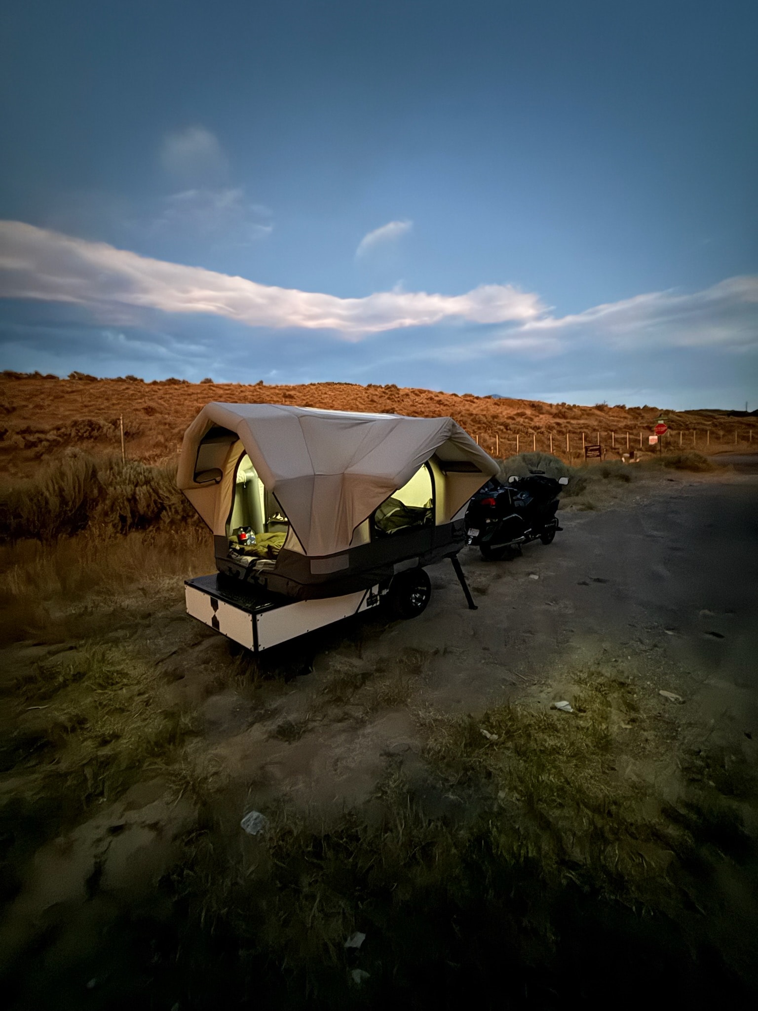 Ultra Compact Conestoga trailer with its airframe tent deployed and glowing at dusk, hitched to a touring motorcycle